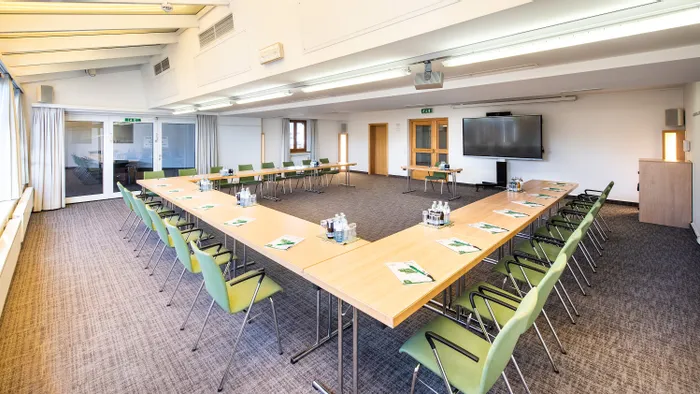Geräumiger Konferenzraum mit U-förmiger Tischanordnung, grünen Stühlen, Wasserflaschen und Präsentationsbildschirm
